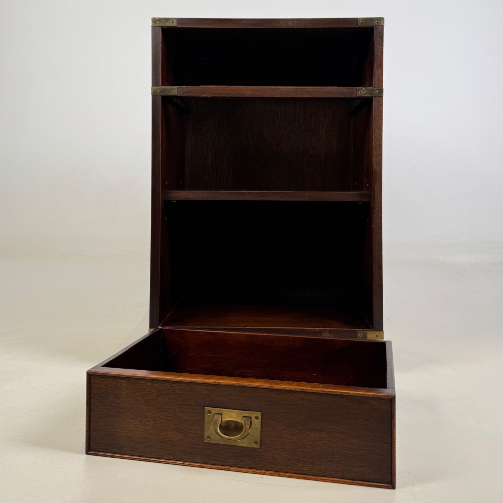 A dark mahogany campaign style cabinet featuring a single upper drawer with a flush brass handle, brass corner mounts, and two open lower shelves. View X
