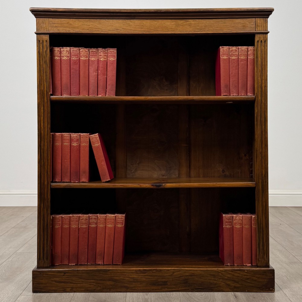 An antique Edwardian open oak bookcase featuring three wide shelves filled with red hardback books, flanked by reeded side pilasters and supported by a solid plinth base.