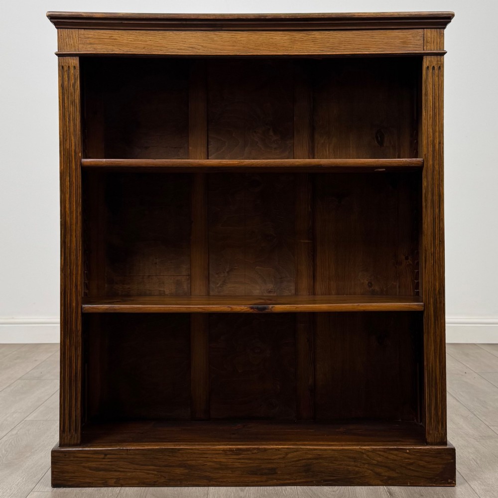 An antique Edwardian open oak bookcase featuring three wide shelves filled with red hardback books, flanked by reeded side pilasters and supported by a solid plinth base. View II