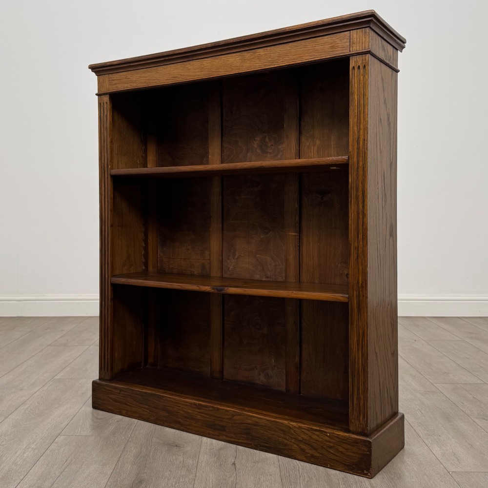 An antique Edwardian open oak bookcase featuring three wide shelves filled with red hardback books, flanked by reeded side pilasters and supported by a solid plinth base. View III