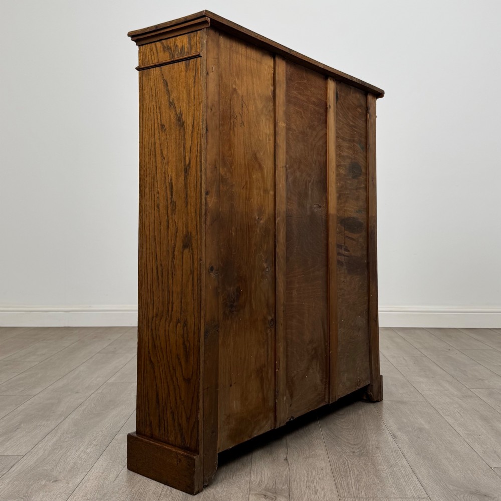 An antique Edwardian open oak bookcase featuring three wide shelves filled with red hardback books, flanked by reeded side pilasters and supported by a solid plinth base. View V