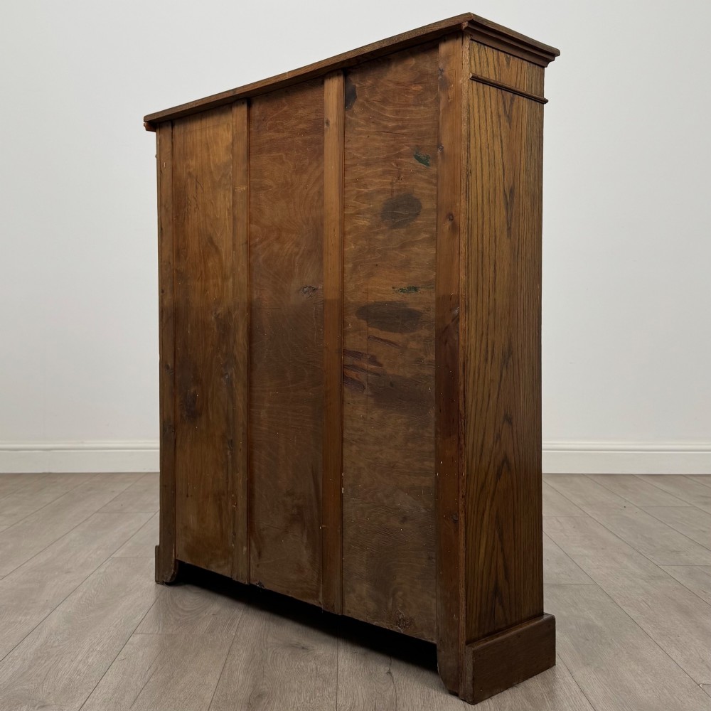 An antique Edwardian open oak bookcase featuring three wide shelves filled with red hardback books, flanked by reeded side pilasters and supported by a solid plinth base. View VII