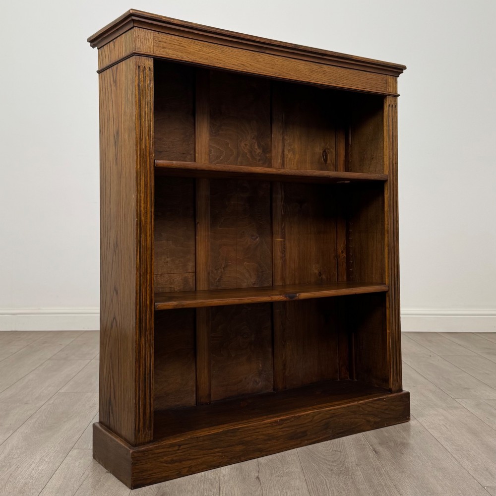 An antique Edwardian open oak bookcase featuring three wide shelves filled with red hardback books, flanked by reeded side pilasters and supported by a solid plinth base. View IX