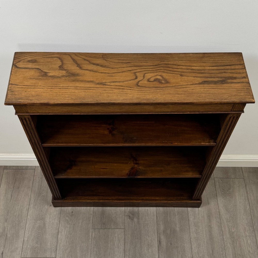 An antique Edwardian open oak bookcase featuring three wide shelves filled with red hardback books, flanked by reeded side pilasters and supported by a solid plinth base. View X