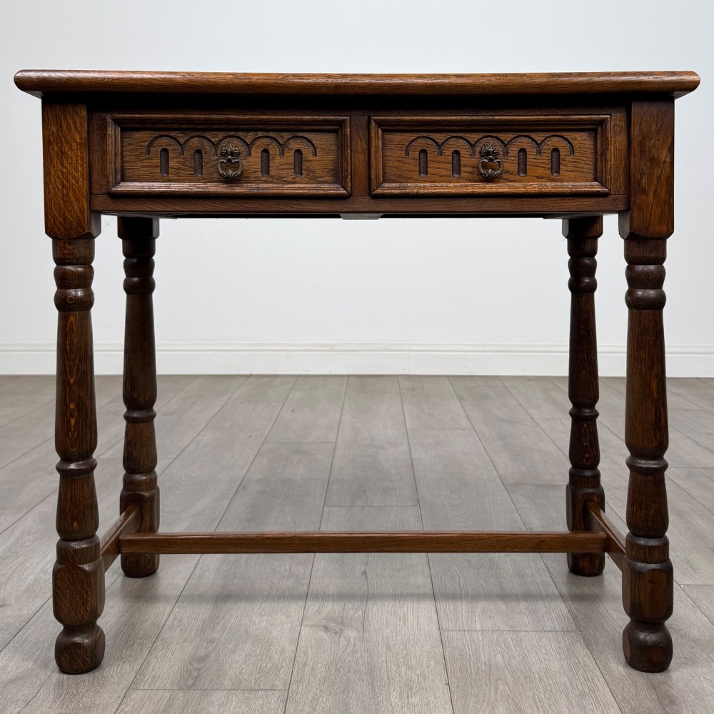 An antique Edwardian dark oak side table featuring two carved front drawers with brass ring pulls and turned legs joined by stretchers.