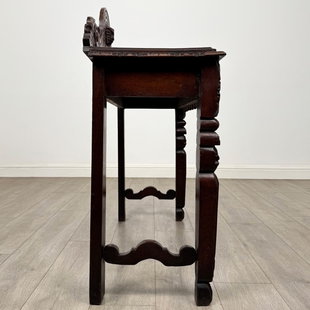 A dark brown 19th-century carved oak side table featuring an ornate splashback with a lion mask carving and intricately carved legs. View VII