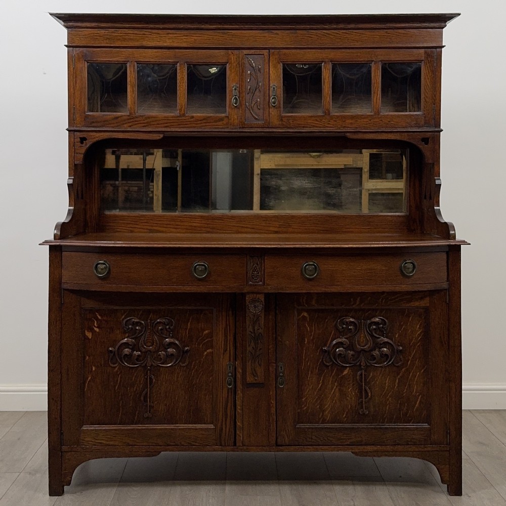 An early twentieth-century Arts and Crafts oak sideboard featuring carved floral door panels, two drawers, a mirrored backsplash, and an upper glazed cabinet.