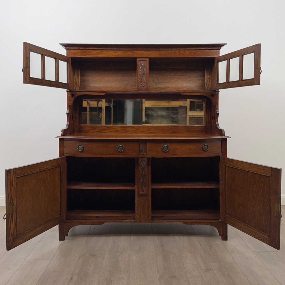 An early twentieth-century Arts and Crafts oak sideboard featuring carved floral door panels, two drawers, a mirrored backsplash, and an upper glazed cabinet. View II