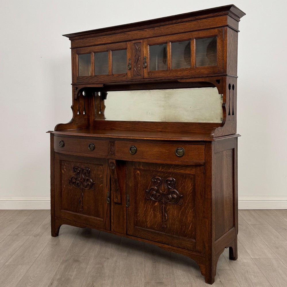 An early twentieth-century Arts and Crafts oak sideboard featuring carved floral door panels, two drawers, a mirrored backsplash, and an upper glazed cabinet. View III