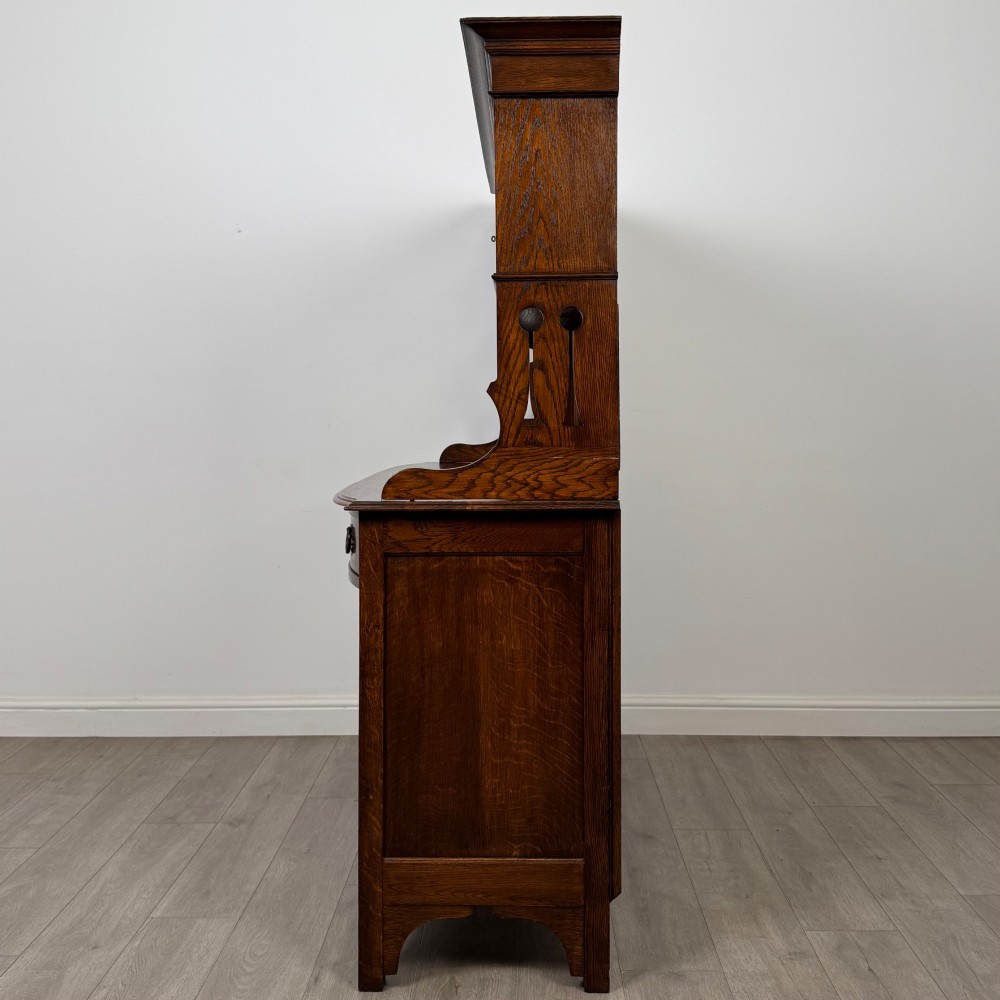 An early twentieth-century Arts and Crafts oak sideboard featuring carved floral door panels, two drawers, a mirrored backsplash, and an upper glazed cabinet. View IV