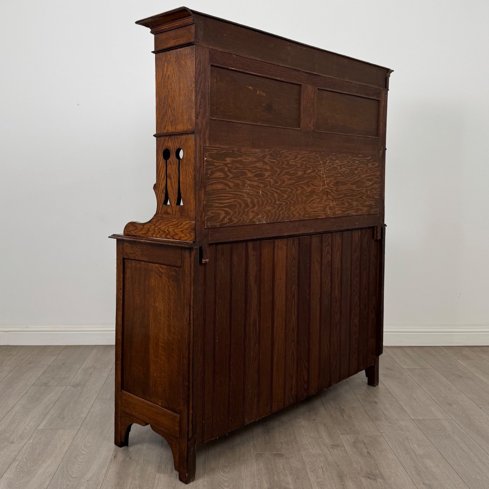 An early twentieth-century Arts and Crafts oak sideboard featuring carved floral door panels, two drawers, a mirrored backsplash, and an upper glazed cabinet. View V