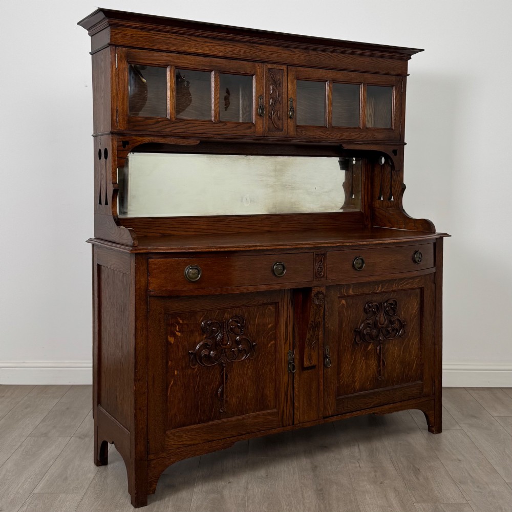 An early twentieth-century Arts and Crafts oak sideboard featuring carved floral door panels, two drawers, a mirrored backsplash, and an upper glazed cabinet. View X