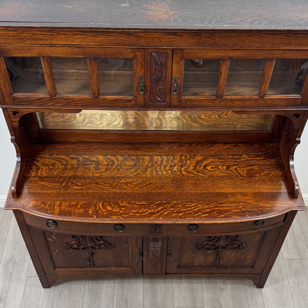 An early twentieth-century Arts and Crafts oak sideboard featuring carved floral door panels, two drawers, a mirrored backsplash, and an upper glazed cabinet. View XI