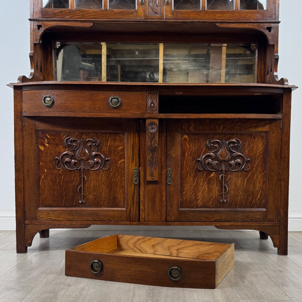 An early twentieth-century Arts and Crafts oak sideboard featuring carved floral door panels, two drawers, a mirrored backsplash, and an upper glazed cabinet. View XII