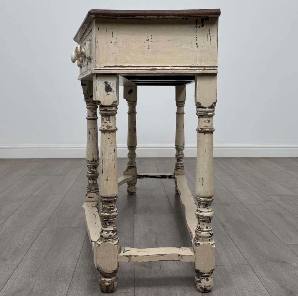 A 20th-century country house style console table featuring a distressed cream-painted carved base with five turned legs and a polished dark oak top. View III