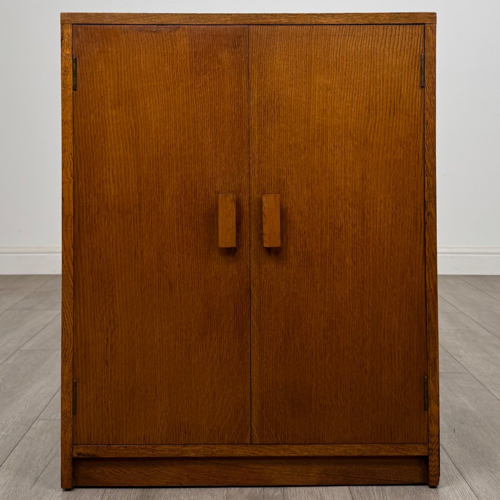 A freestanding golden oak cupboard featuring two flush rectangular doors with matching wooden block handles and brass hinges.