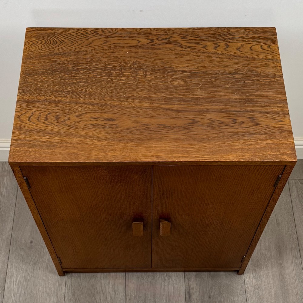 A freestanding golden oak cupboard featuring two flush rectangular doors with matching wooden block handles and brass hinges. View X