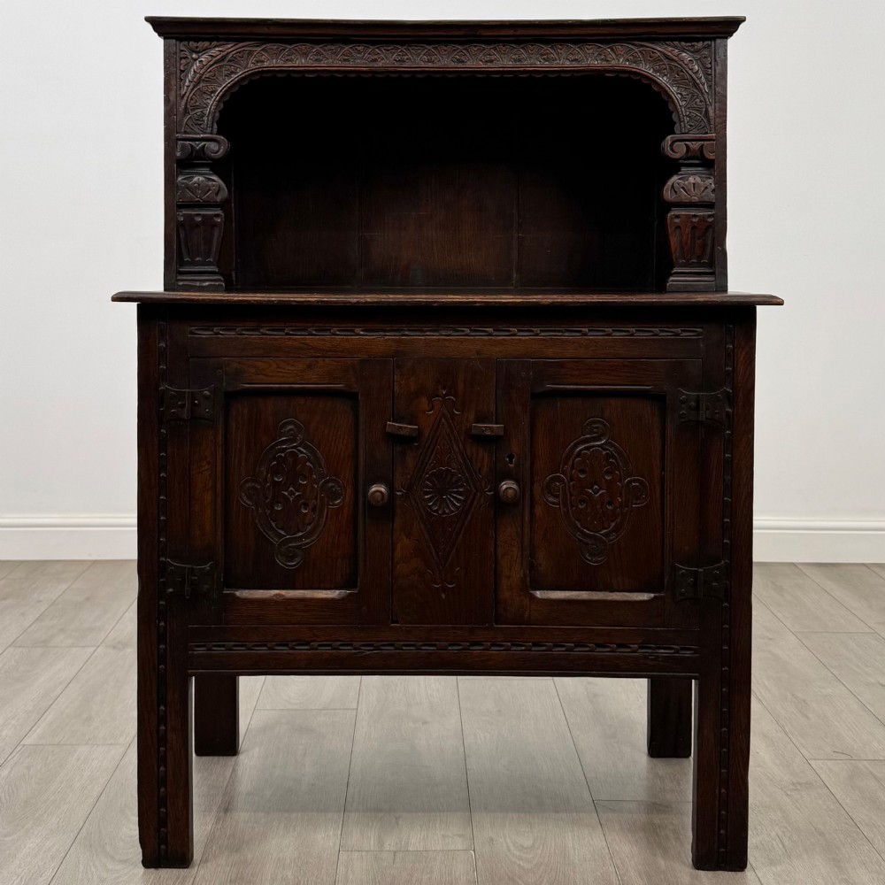 A dark brown 19th-century carved oak cupboard featuring an open upper shelf supported by carved pillars, above a lower two-door cabinet with relief-carved panels and iron hinges.