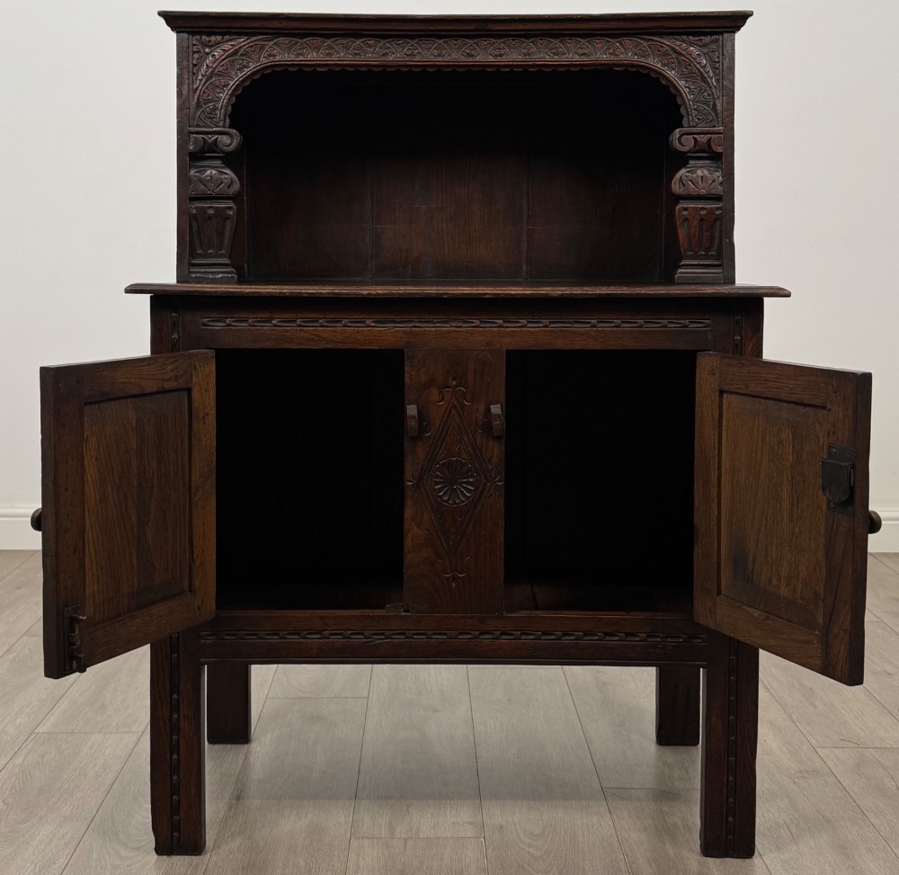A dark brown 19th-century carved oak cupboard featuring an open upper shelf supported by carved pillars, above a lower two-door cabinet with relief-carved panels and iron hinges. View II