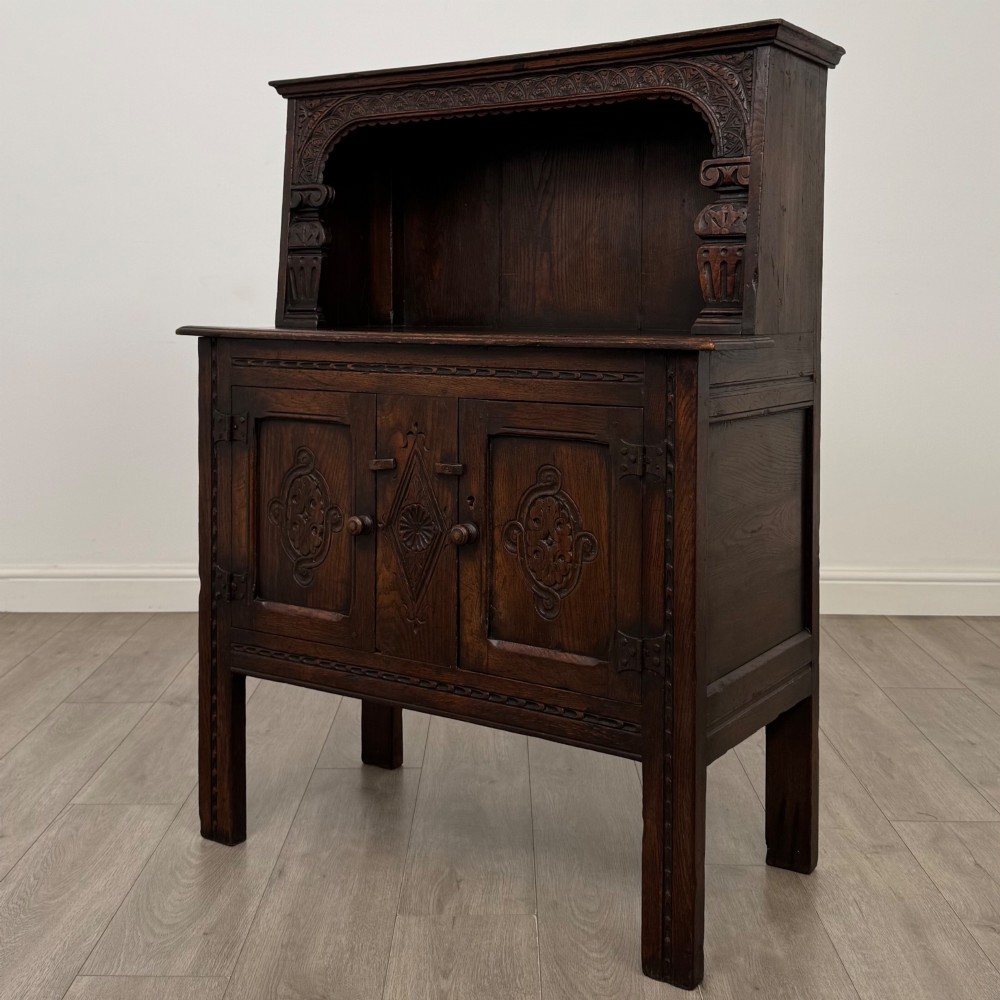 A dark brown 19th-century carved oak cupboard featuring an open upper shelf supported by carved pillars, above a lower two-door cabinet with relief-carved panels and iron hinges. View III