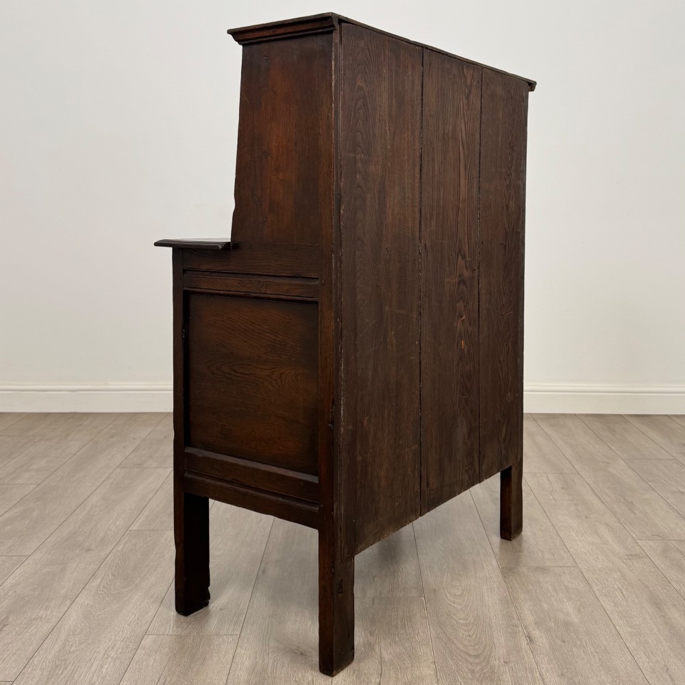 A dark brown 19th-century carved oak cupboard featuring an open upper shelf supported by carved pillars, above a lower two-door cabinet with relief-carved panels and iron hinges. View V