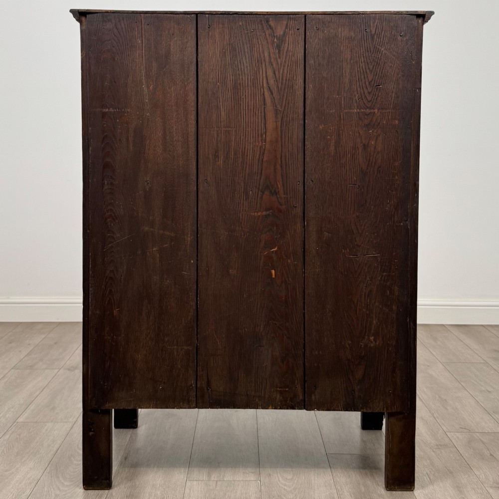 A dark brown 19th-century carved oak cupboard featuring an open upper shelf supported by carved pillars, above a lower two-door cabinet with relief-carved panels and iron hinges. View VI