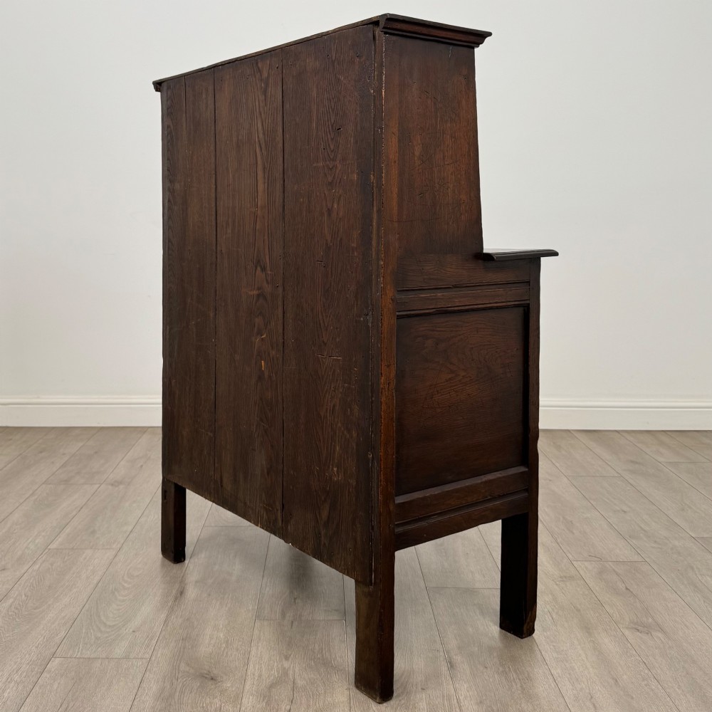 A dark brown 19th-century carved oak cupboard featuring an open upper shelf supported by carved pillars, above a lower two-door cabinet with relief-carved panels and iron hinges. View VII