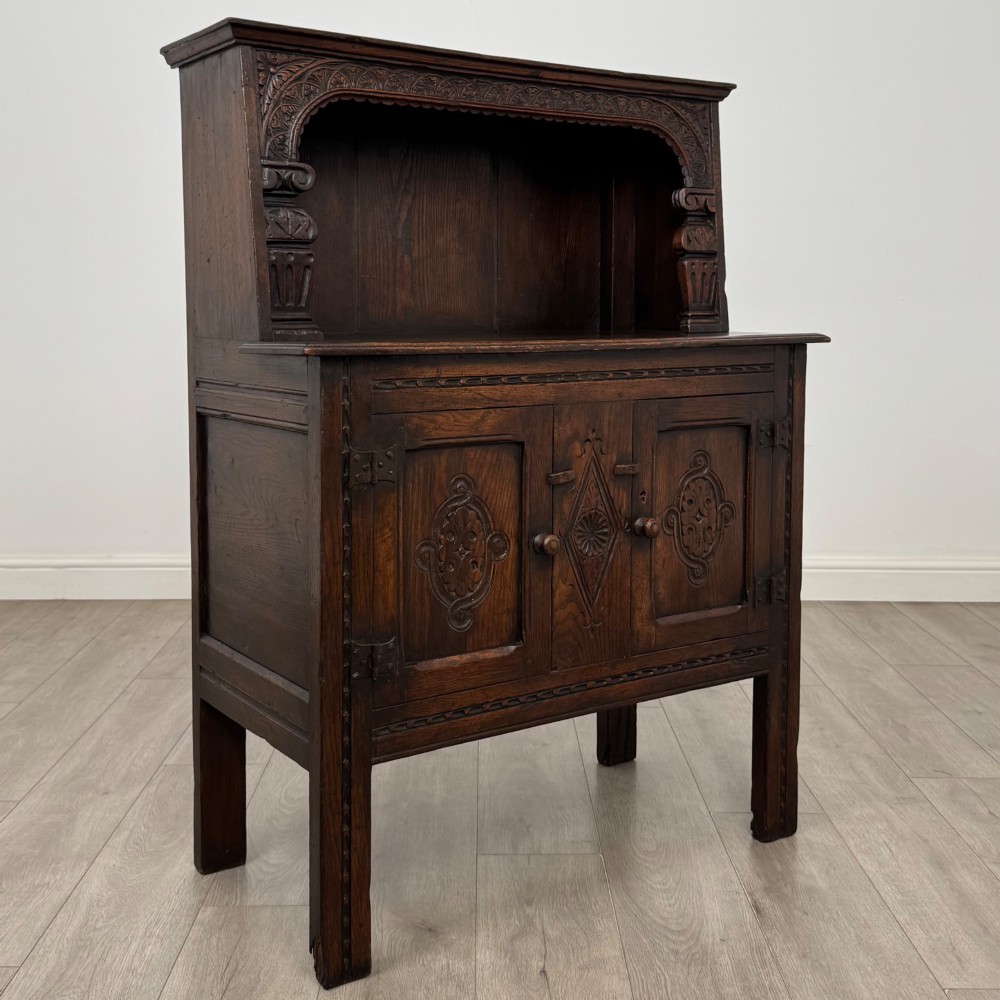 A dark brown 19th-century carved oak cupboard featuring an open upper shelf supported by carved pillars, above a lower two-door cabinet with relief-carved panels and iron hinges. View IX