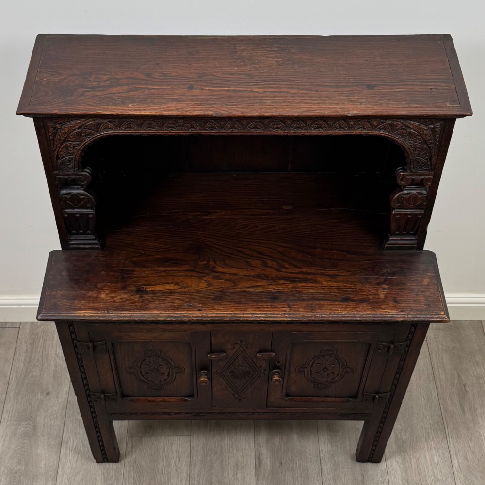 A dark brown 19th-century carved oak cupboard featuring an open upper shelf supported by carved pillars, above a lower two-door cabinet with relief-carved panels and iron hinges. View X