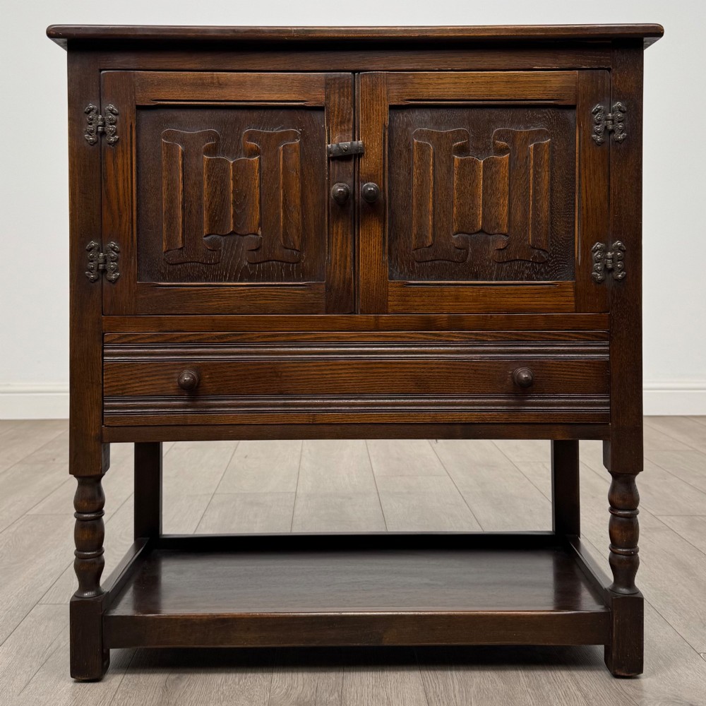 A dark oak cupboard featuring two linenfold carved doors, iron butterfly hinges, a single lower drawer, turned legs, and a bottom shelf.