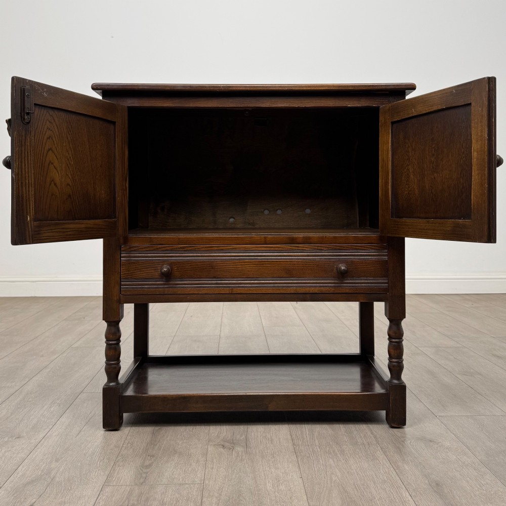 A dark oak cupboard featuring two linenfold carved doors, iron butterfly hinges, a single lower drawer, turned legs, and a bottom shelf. View II