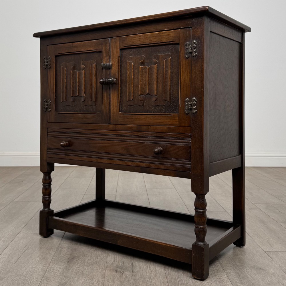 A dark oak cupboard featuring two linenfold carved doors, iron butterfly hinges, a single lower drawer, turned legs, and a bottom shelf. View III