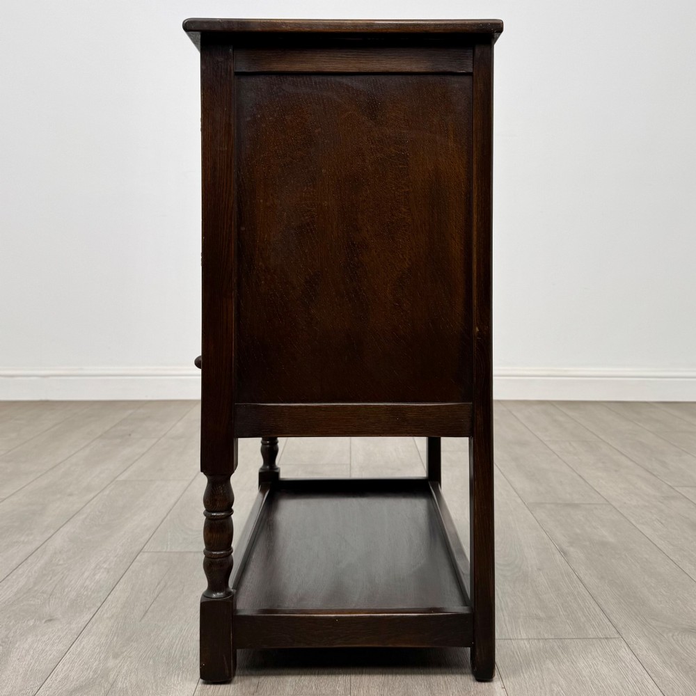 A dark oak cupboard featuring two linenfold carved doors, iron butterfly hinges, a single lower drawer, turned legs, and a bottom shelf. View IV