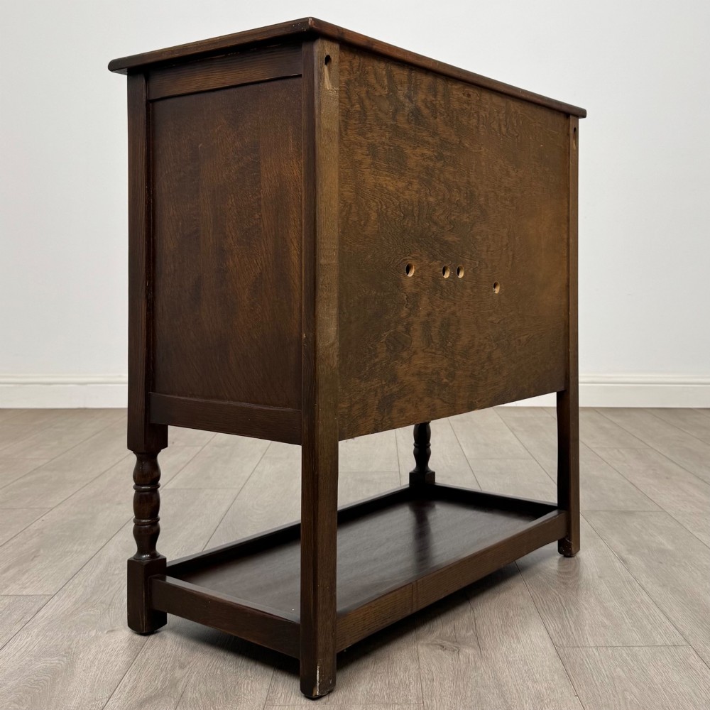 A dark oak cupboard featuring two linenfold carved doors, iron butterfly hinges, a single lower drawer, turned legs, and a bottom shelf. View V