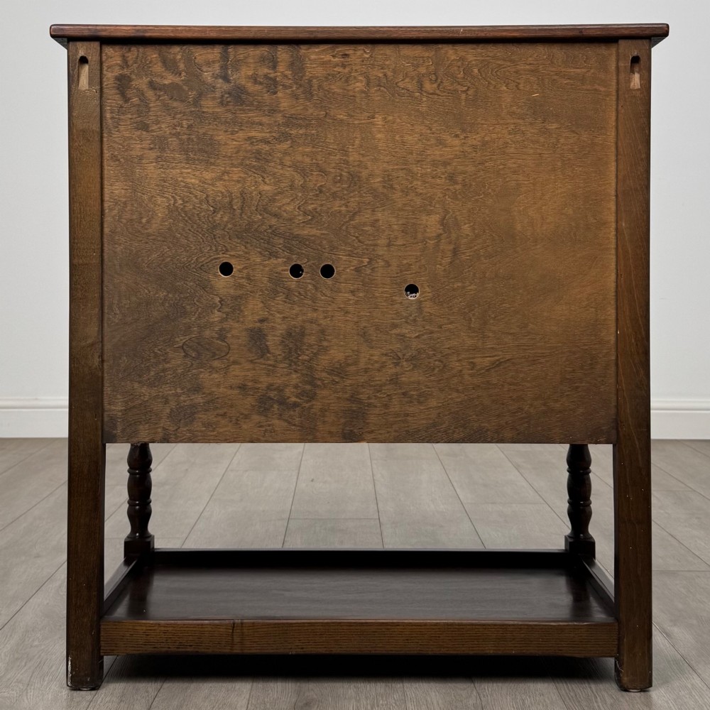 A dark oak cupboard featuring two linenfold carved doors, iron butterfly hinges, a single lower drawer, turned legs, and a bottom shelf. View VI