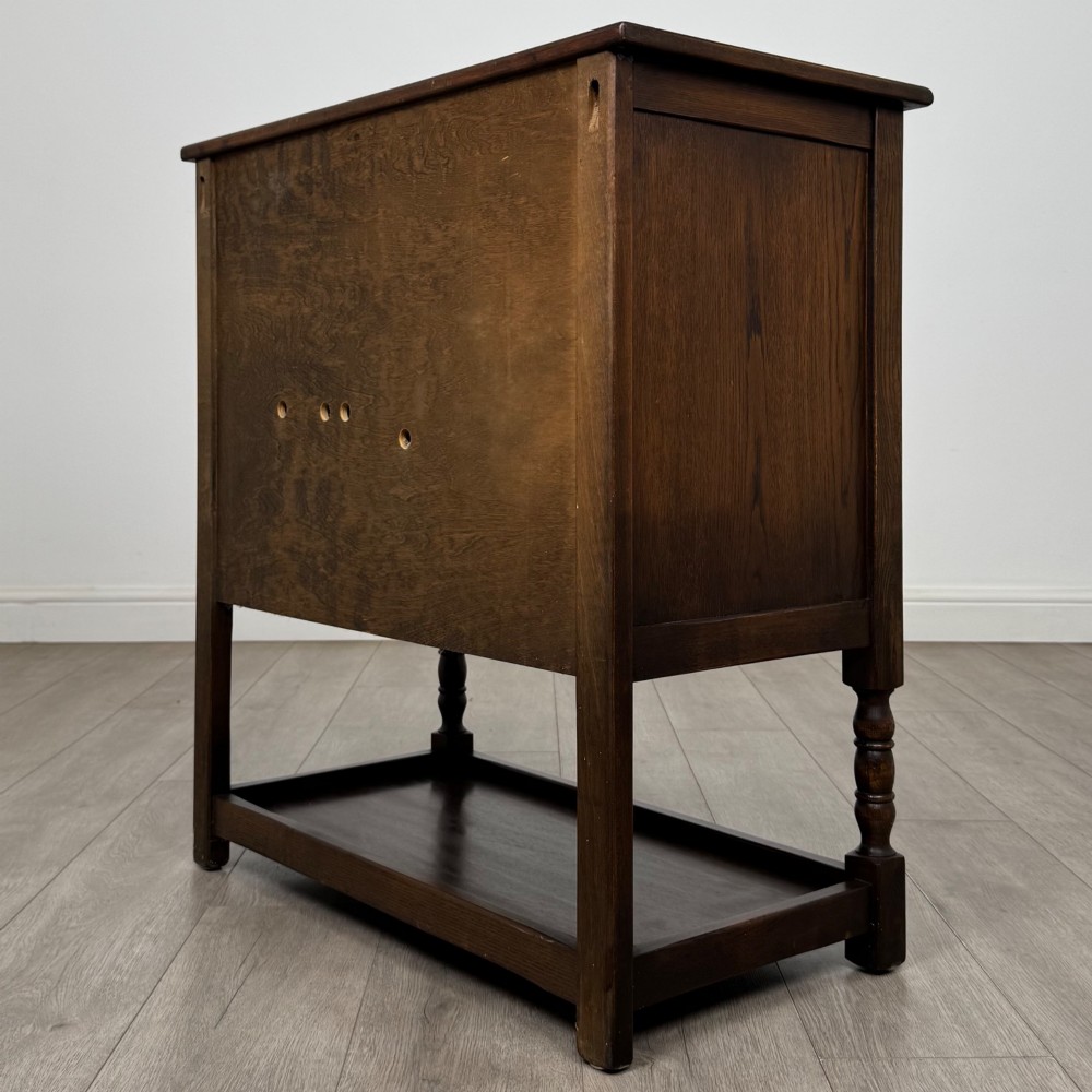 A dark oak cupboard featuring two linenfold carved doors, iron butterfly hinges, a single lower drawer, turned legs, and a bottom shelf. View VII