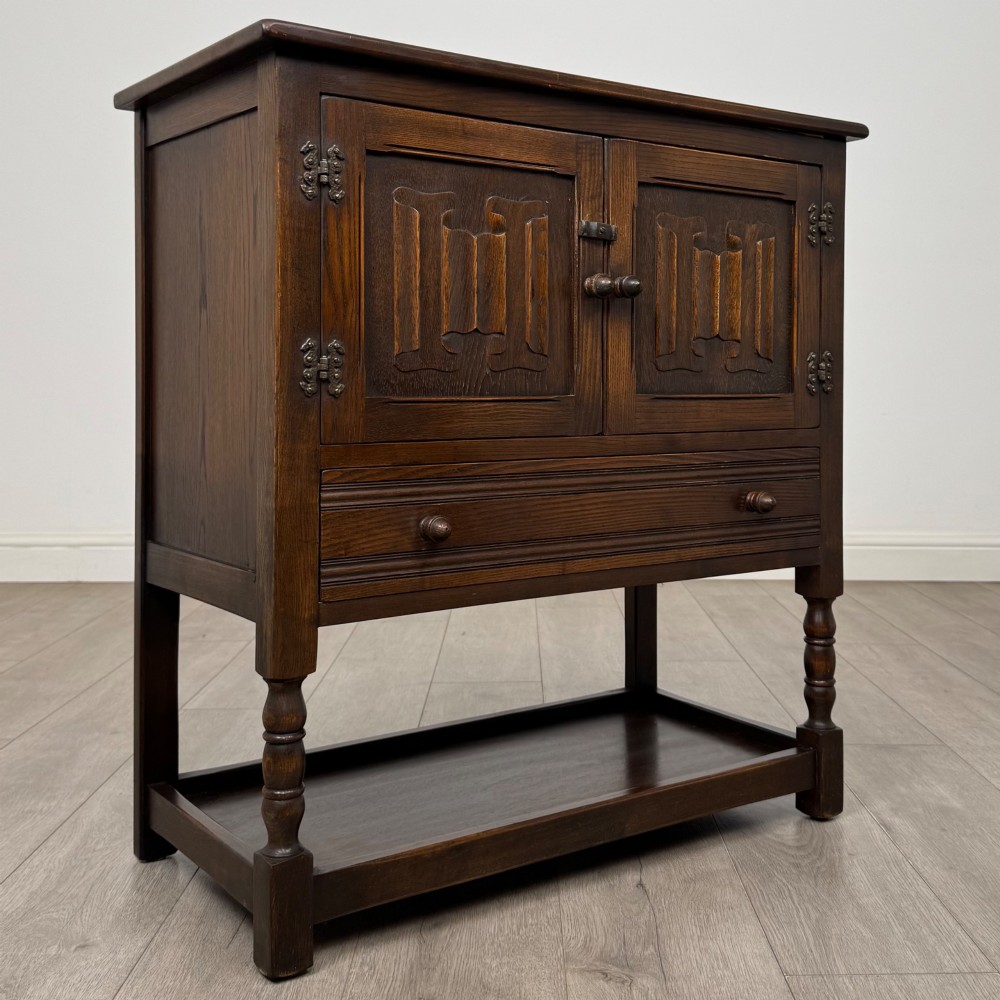 A dark oak cupboard featuring two linenfold carved doors, iron butterfly hinges, a single lower drawer, turned legs, and a bottom shelf. View IX