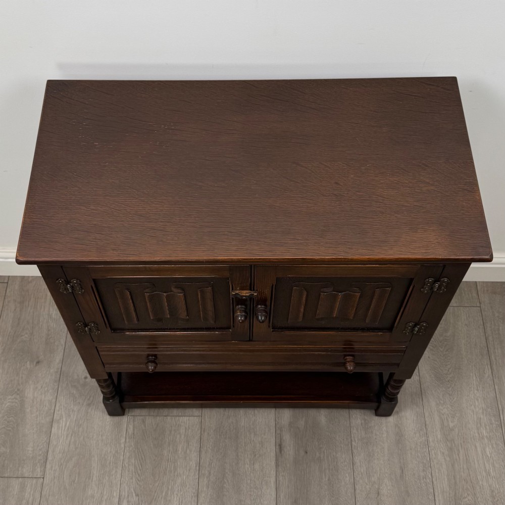 A dark oak cupboard featuring two linenfold carved doors, iron butterfly hinges, a single lower drawer, turned legs, and a bottom shelf. View X