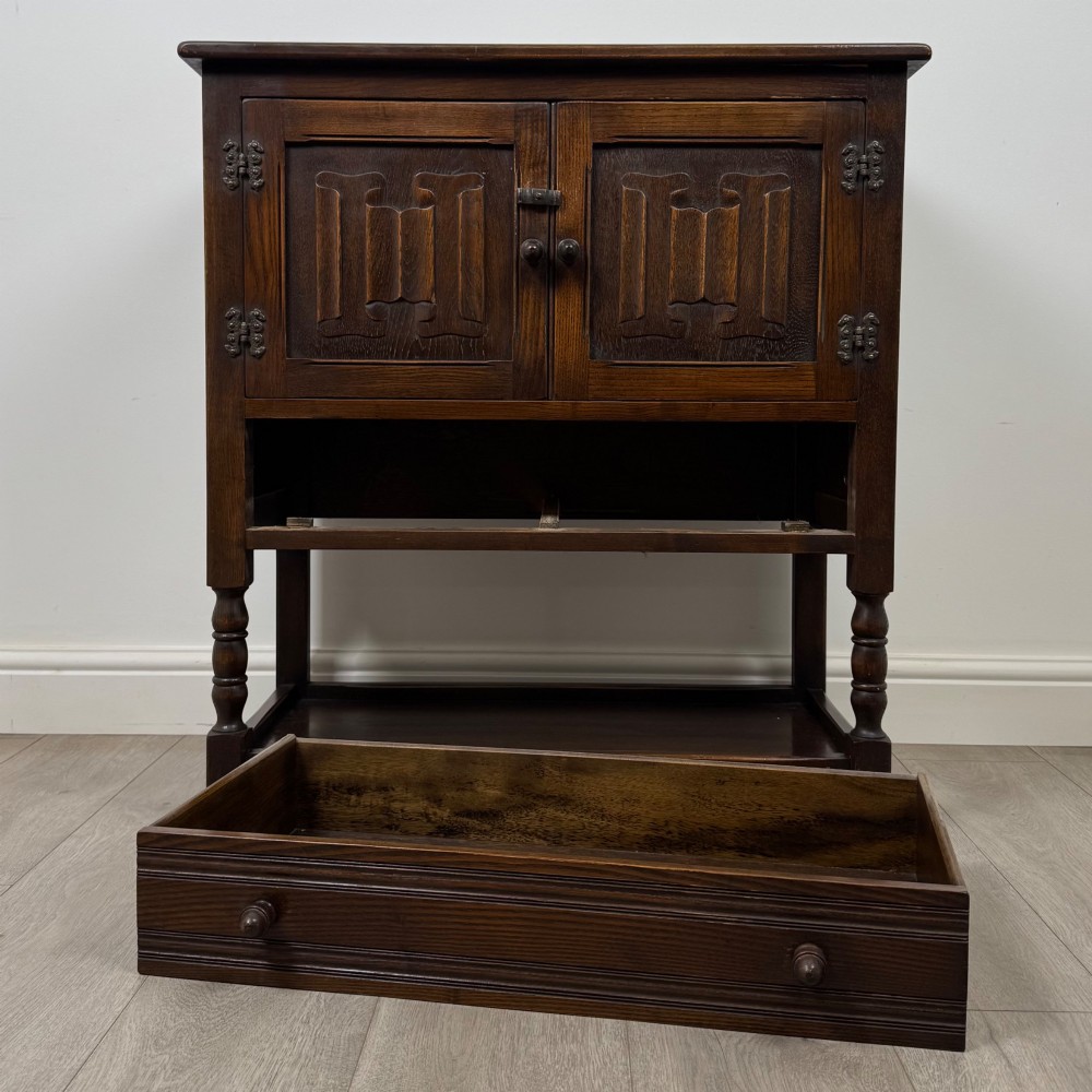 A dark oak cupboard featuring two linenfold carved doors, iron butterfly hinges, a single lower drawer, turned legs, and a bottom shelf. View XI