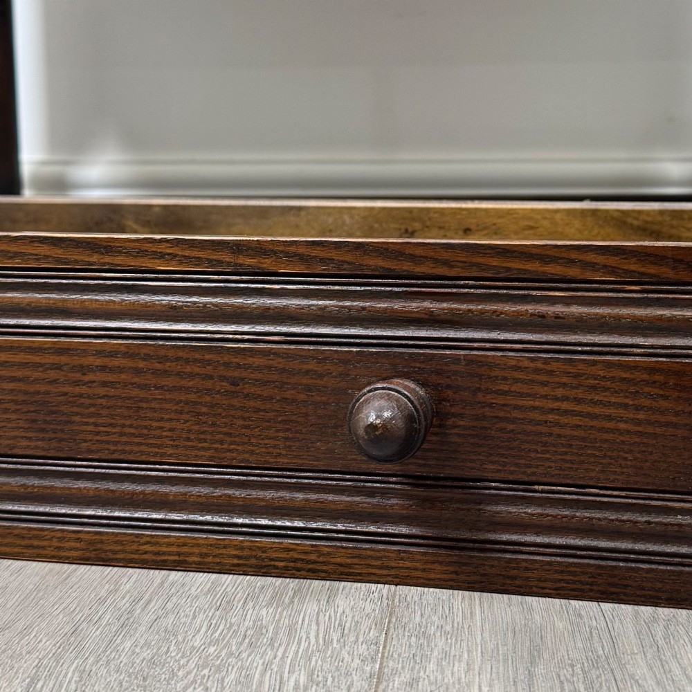 A dark oak cupboard featuring two linenfold carved doors, iron butterfly hinges, a single lower drawer, turned legs, and a bottom shelf. View XII