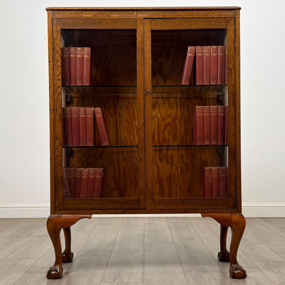 An antique 20th-century oak display cabinet featuring twin glazed doors, glass interior shelves, a carved top frieze, and cabriole legs ending in ball-and-claw feet.