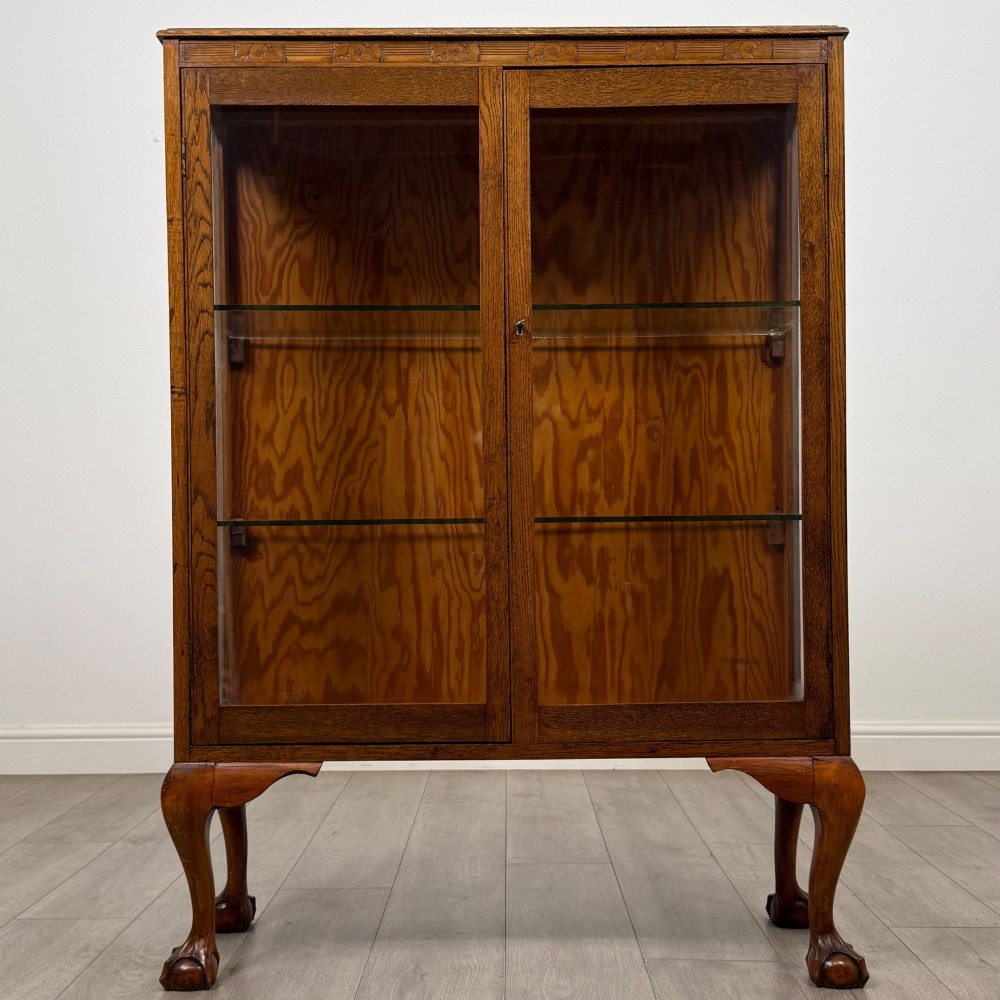 An antique 20th-century oak display cabinet featuring twin glazed doors, glass interior shelves, a carved top frieze, and cabriole legs ending in ball-and-claw feet. View II