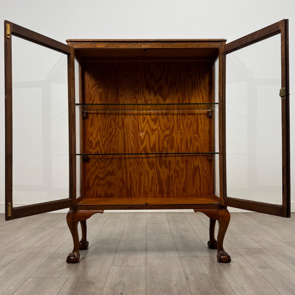 An antique 20th-century oak display cabinet featuring twin glazed doors, glass interior shelves, a carved top frieze, and cabriole legs ending in ball-and-claw feet. View III
