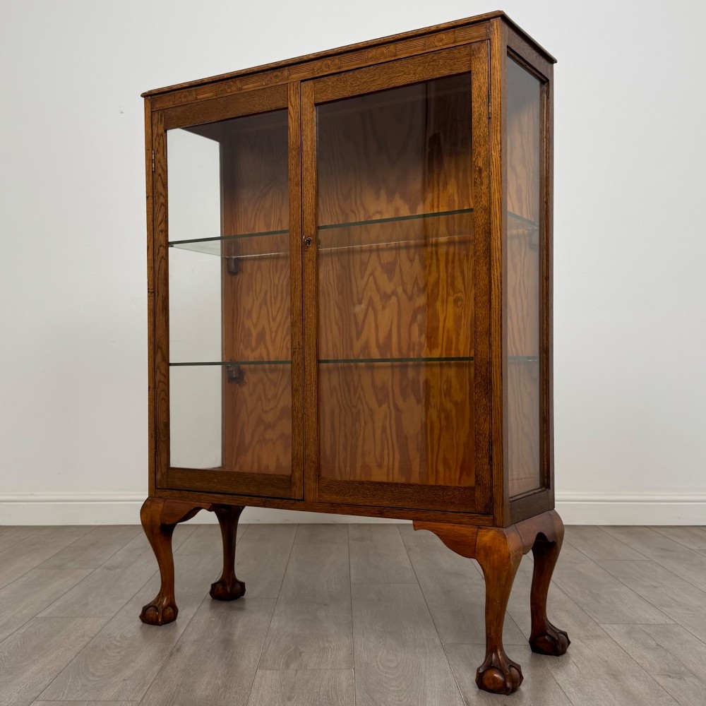 An antique 20th-century oak display cabinet featuring twin glazed doors, glass interior shelves, a carved top frieze, and cabriole legs ending in ball-and-claw feet. View IV