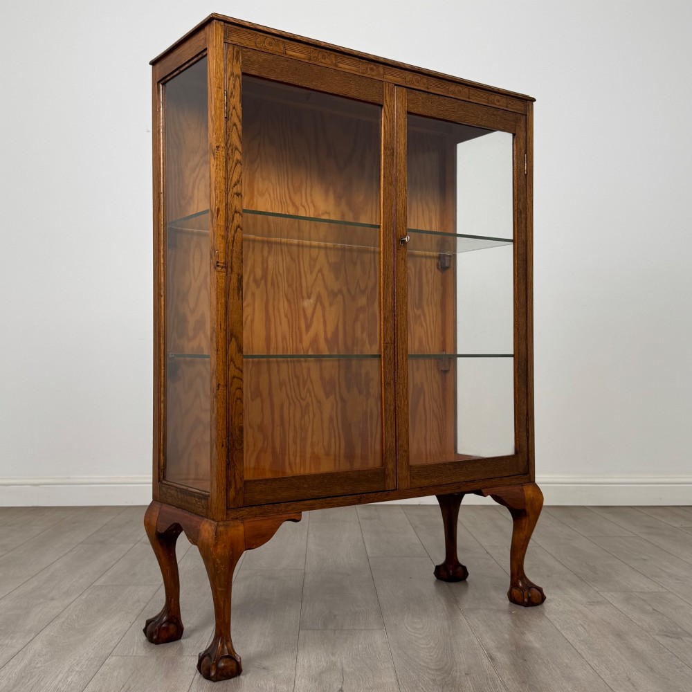 An antique 20th-century oak display cabinet featuring twin glazed doors, glass interior shelves, a carved top frieze, and cabriole legs ending in ball-and-claw feet. View X