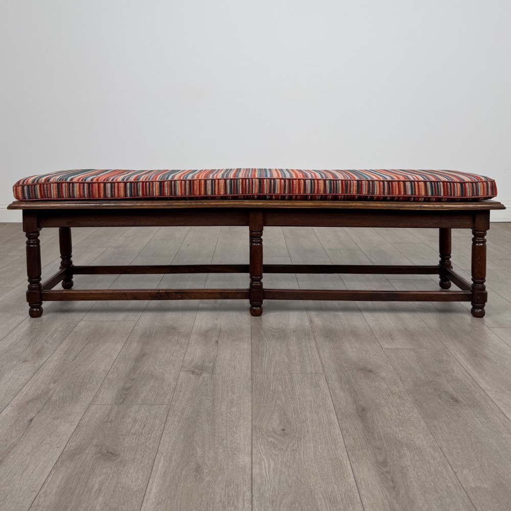 A long dark oak bench featuring six turned legs, perimeter stretchers, and a multi-coloured striped upholstered seat cushion.