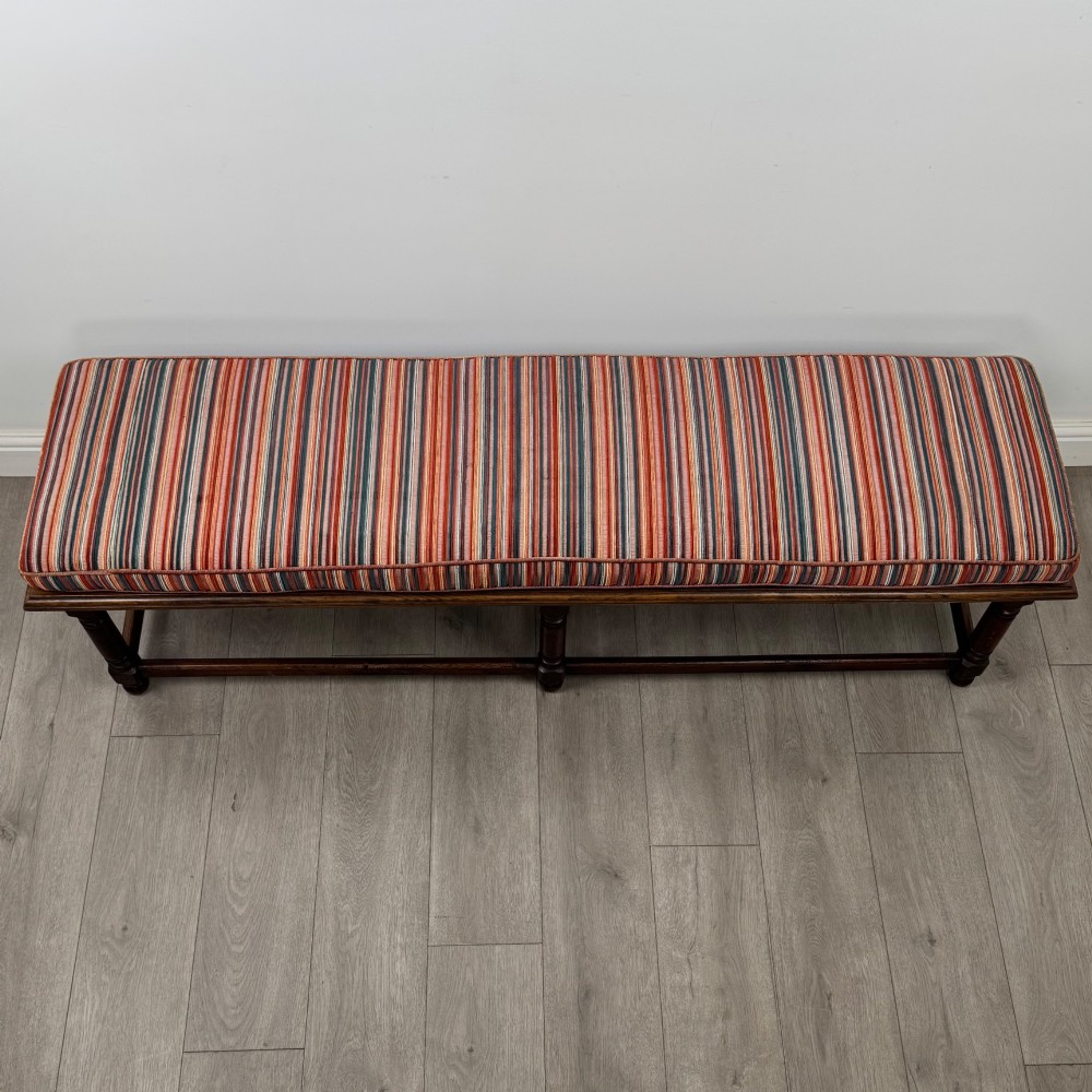 A long dark oak bench featuring six turned legs, perimeter stretchers, and a multi-coloured striped upholstered seat cushion. View X