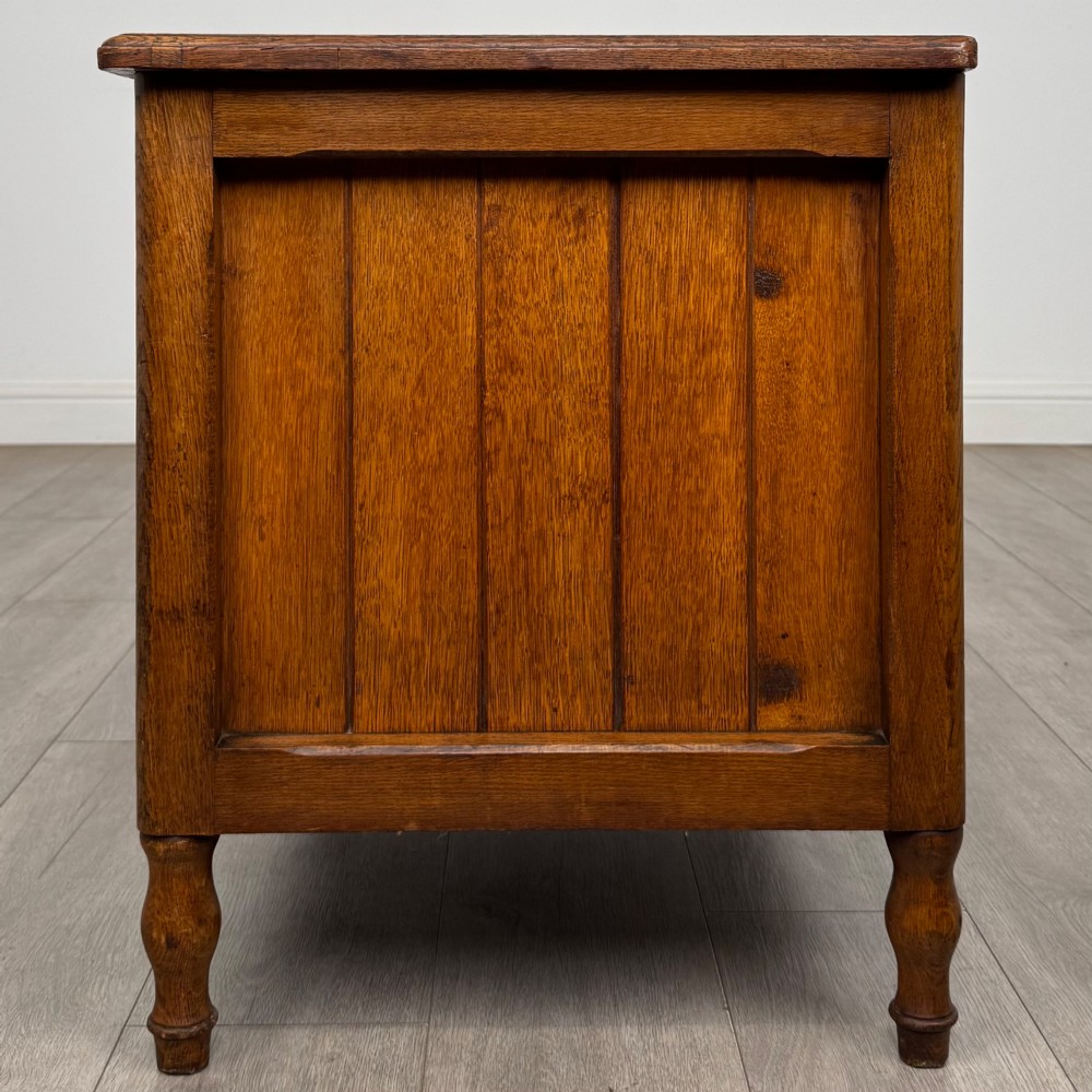 A golden oak Arts and Crafts blanket box featuring vertical panelled sides, a central escutcheon keyhole, and turned legs. View III