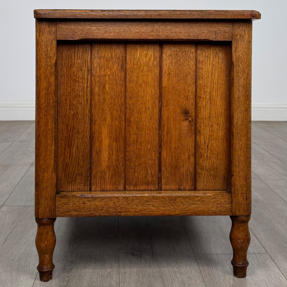 A golden oak Arts and Crafts blanket box featuring vertical panelled sides, a central escutcheon keyhole, and turned legs. View VII