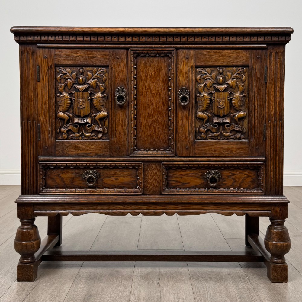 A dark oak cupboard featuring two relief-carved heraldic door panels above two drawers, supported by turned legs and stretchers.